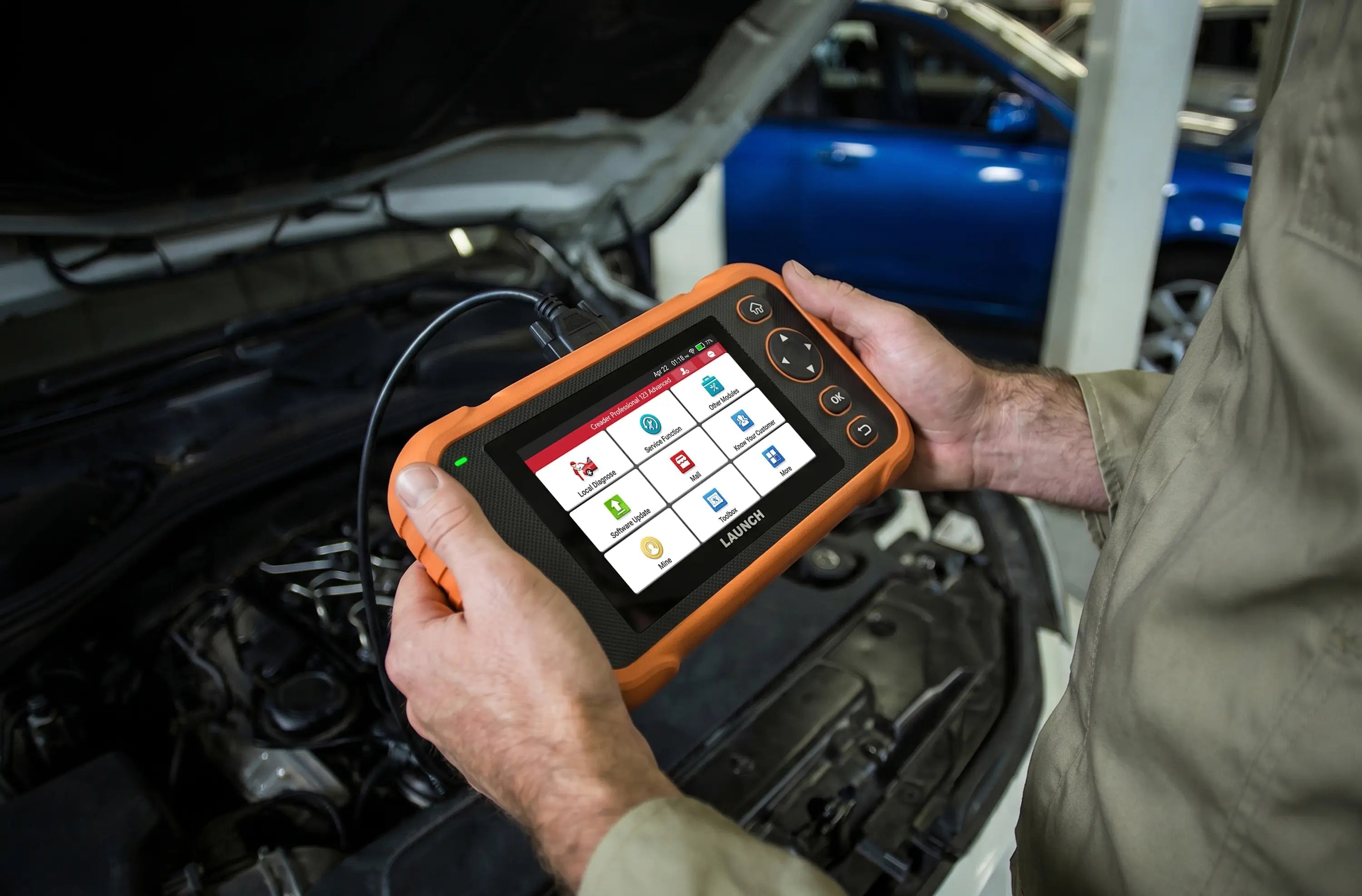 a person's hands holding a LAUNCH CRP123 Advanced diagnostic scanner over an open car engine bay in a garage setting. The device is enclosed in a rugged orange casing and features a color touchscreen displaying various automotive service menu icons.