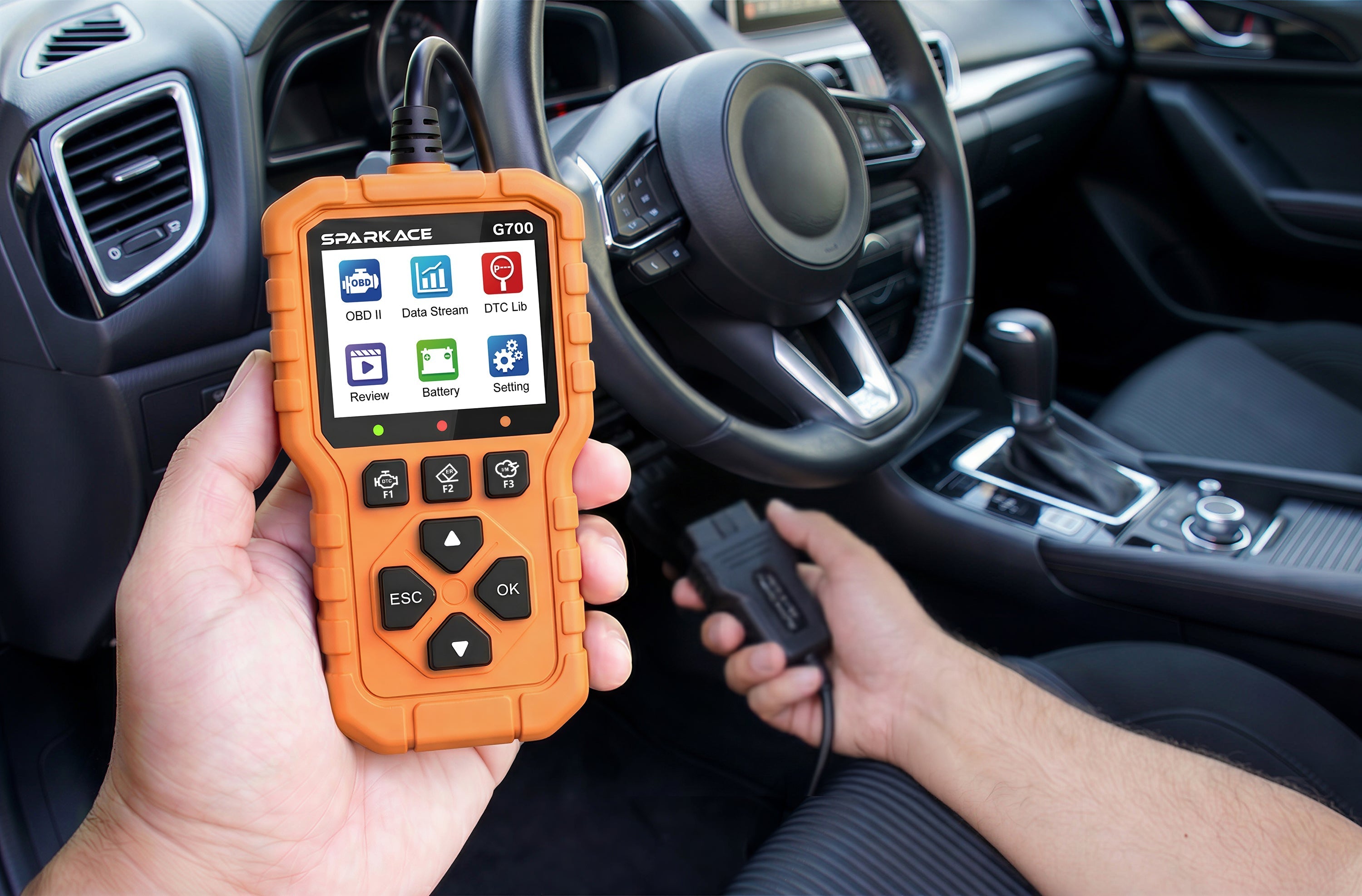 A person's hands are holding an orange OBD II scanner. The device features a color display with menu icons and physical navigation buttons, while the other hand holds the OBD II connector ready to be plugged into the vehicle near the steering wheel area.