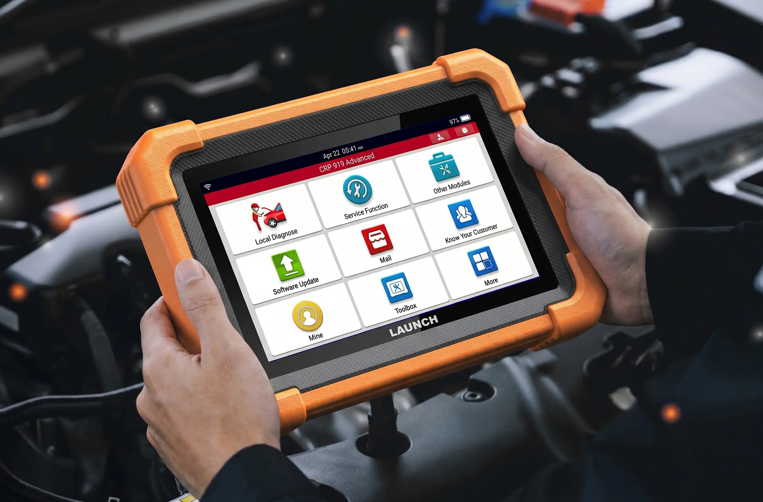 A person holds a LAUNCH CRP 919 Advanced diagnostic scanner over an open car engine bay. The device is enclosed in a rugged orange protective bumper and features a touchscreen displaying various automotive diagnostic menu icons.
