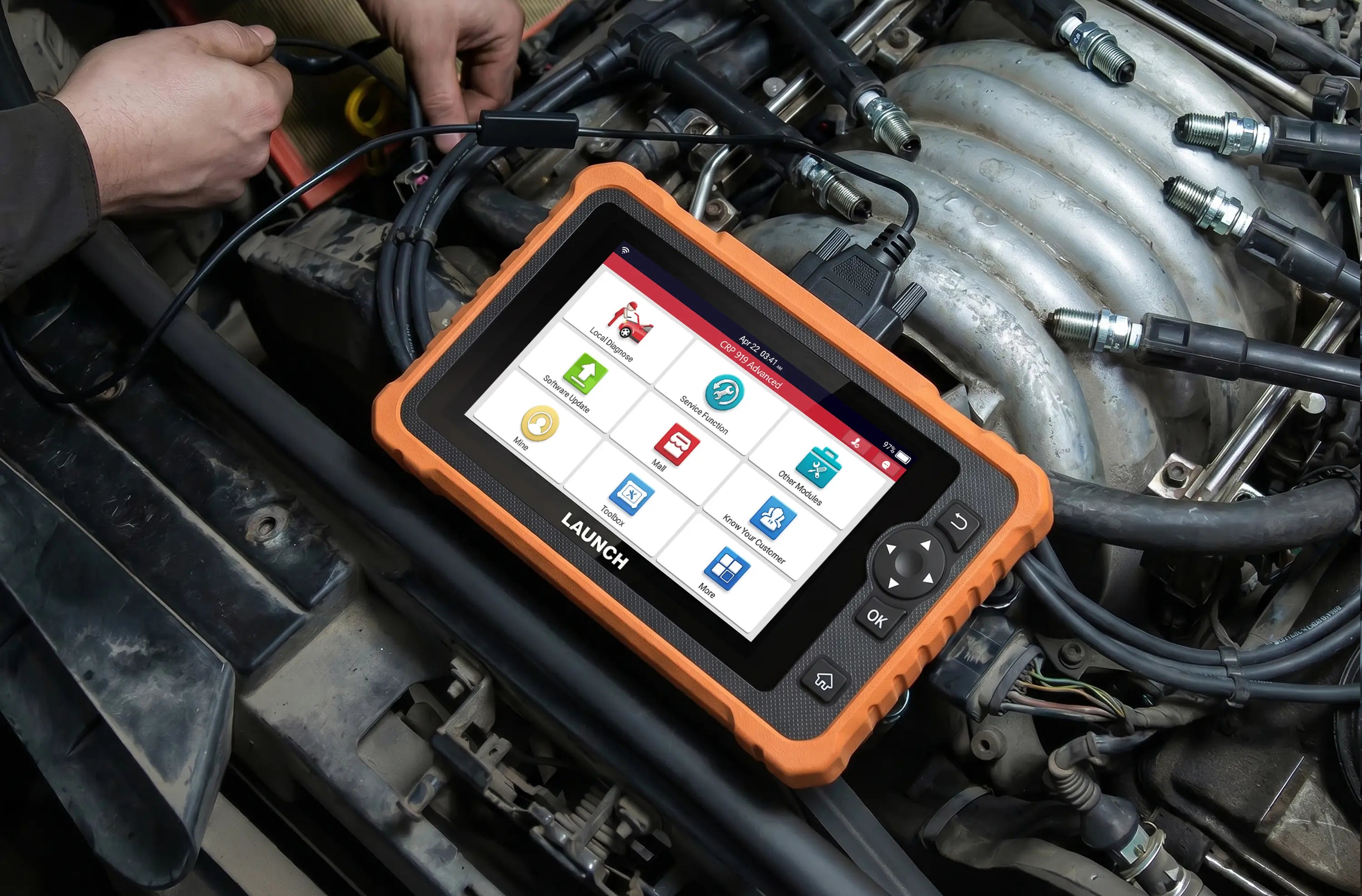A LAUNCH CRP 919 Advanced diagnostic tool resting on an open car engine during a maintenance check. Features a rugged orange casing, a color touchscreen displaying various menu options, and is connected to the vehicle via a black cable.